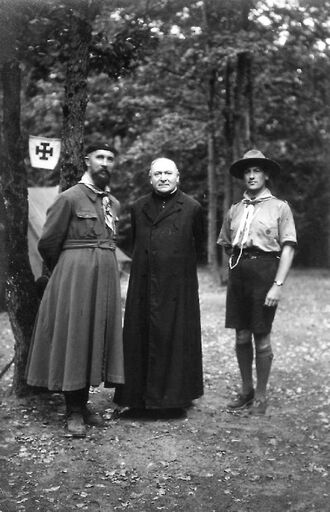 Photo en noir et blanc de trois hommes posant devant quelques arbres : les deux premiers portant une soutane, le troisième est habillé en scout