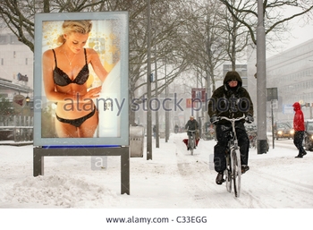 man-cycling-in-harsh-snow-winter-weather-blizzard-while-woman-in-bikini-C33EGG