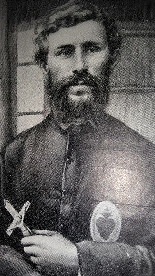 Photo d'un prêtre, barbu, un Sacré-Cœur sur la poitrine et un crucifix à la main, devant une cloison