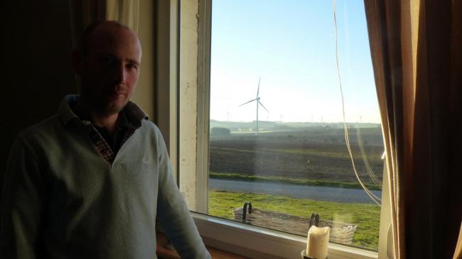 Thibaud Sailly dans sa cuisine avec, en fond, les éoliennes qui jouent avec le soleil.