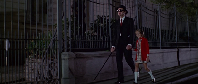 Un homme aveugle et sa petite nièce, vêtue de rouge, marchent mains dans la mains dans une ville nocturne