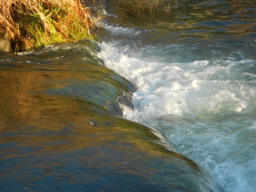 l'eau d'une rivière saute une petite barre de pierres et tombe en écume blanche, rayon de soleil sur l'ensemble