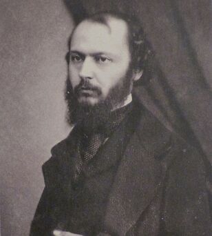 Photographie en noir et blanc d'un homme barbu posant debout, la main gauche sur le ventre.