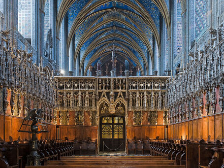 Photo couleur des sièges en bois en deux rangées face à face enserrées dans un écrin de pierre ciselée, au cœur d'une église dont les voutes sont peintes majoritairement de bleu.