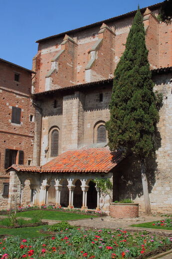 cloître avec colonade romane devant une église en pierre dont la partie supérieure est bâtie en brique de terre cuite rouge.