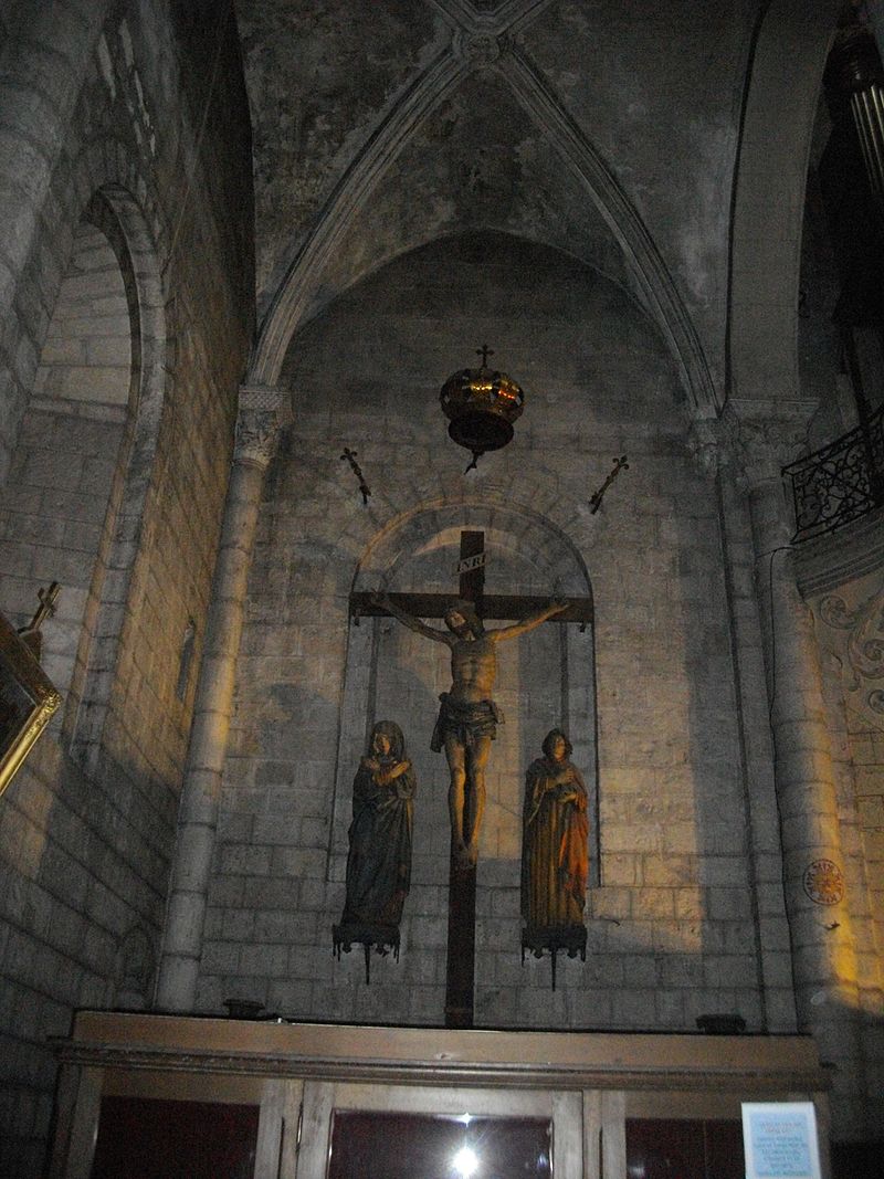 Photo couleur d'un Christ en croix encadré de femmes en prière. La croix est fixés dans une alcôve du mur à linteau roman.